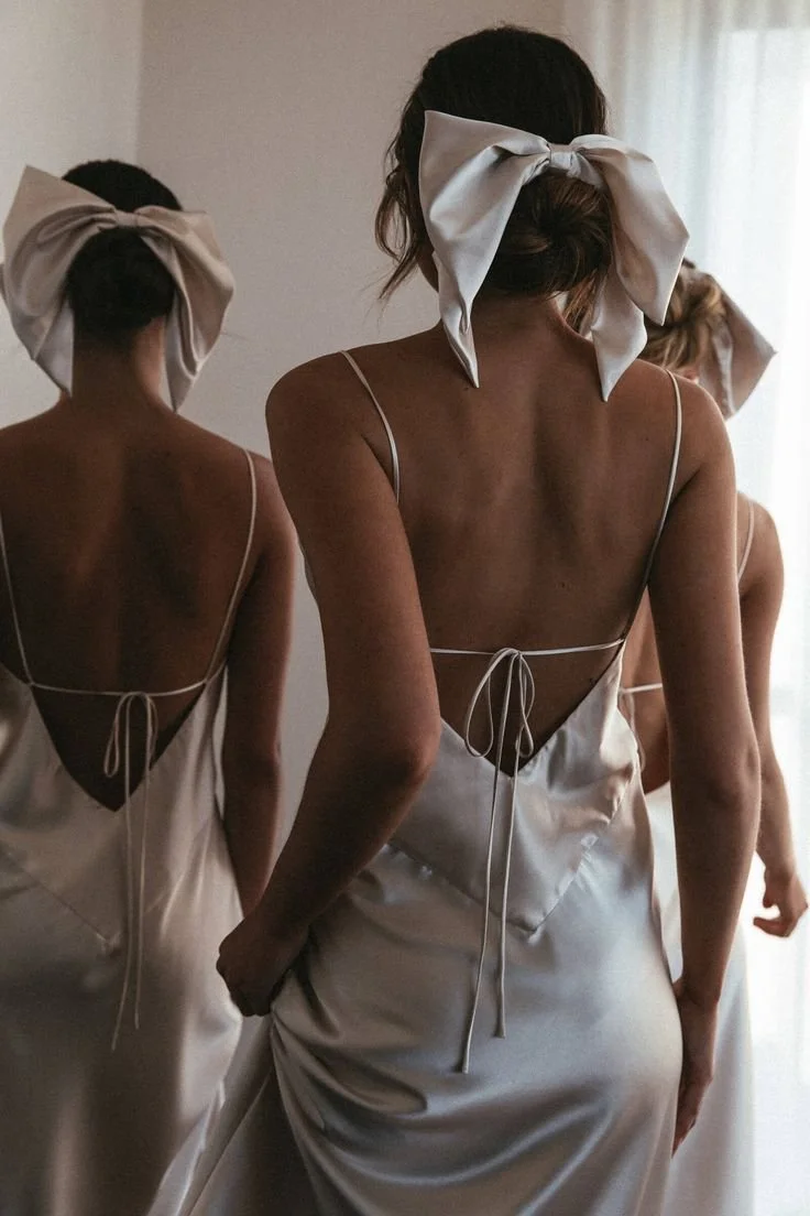 Three women wearing ivory satin dresses with open back designs, tying or adjusting their dress straps, and large satin bows on their heads