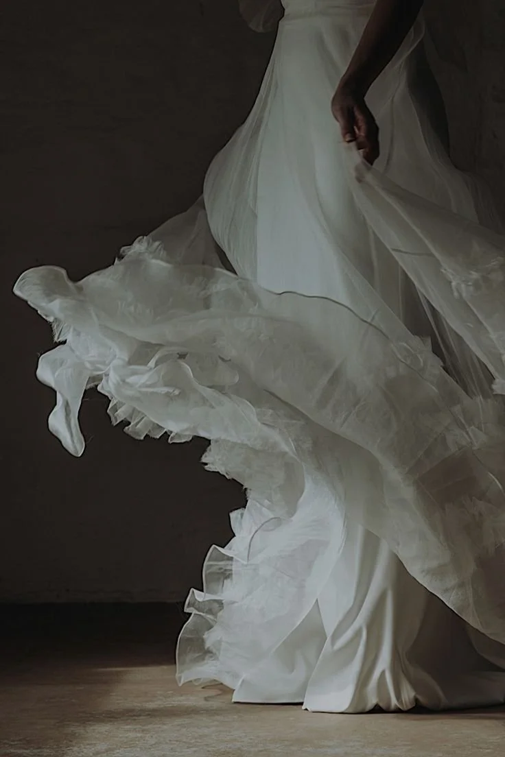 Close-up of a woman in a flowing white wedding dress, partially turned, with her hand gently holding the dress, against a dark background.
