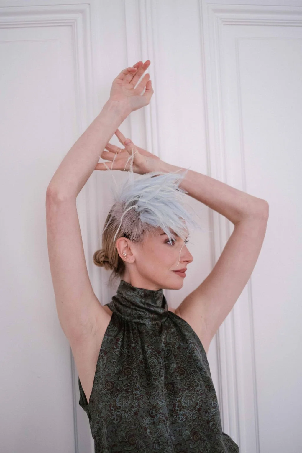 A woman with short, asymmetrical, platinum hair styled in a messy, spiked fashion, posing with her arms raised against a white wall with paneling, wearing a dark, patterned sleeveless top.