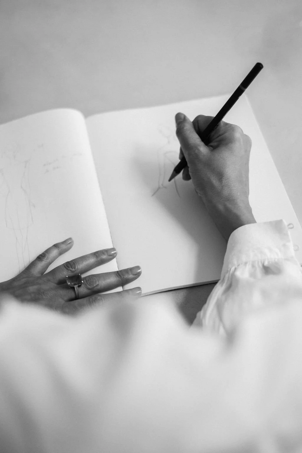 A person drawing in a sketchbook with a pen, with their left hand resting on the open pages. The person is wearing a long-sleeve shirt, and the sketchbook contains light sketches or sketches in progress.