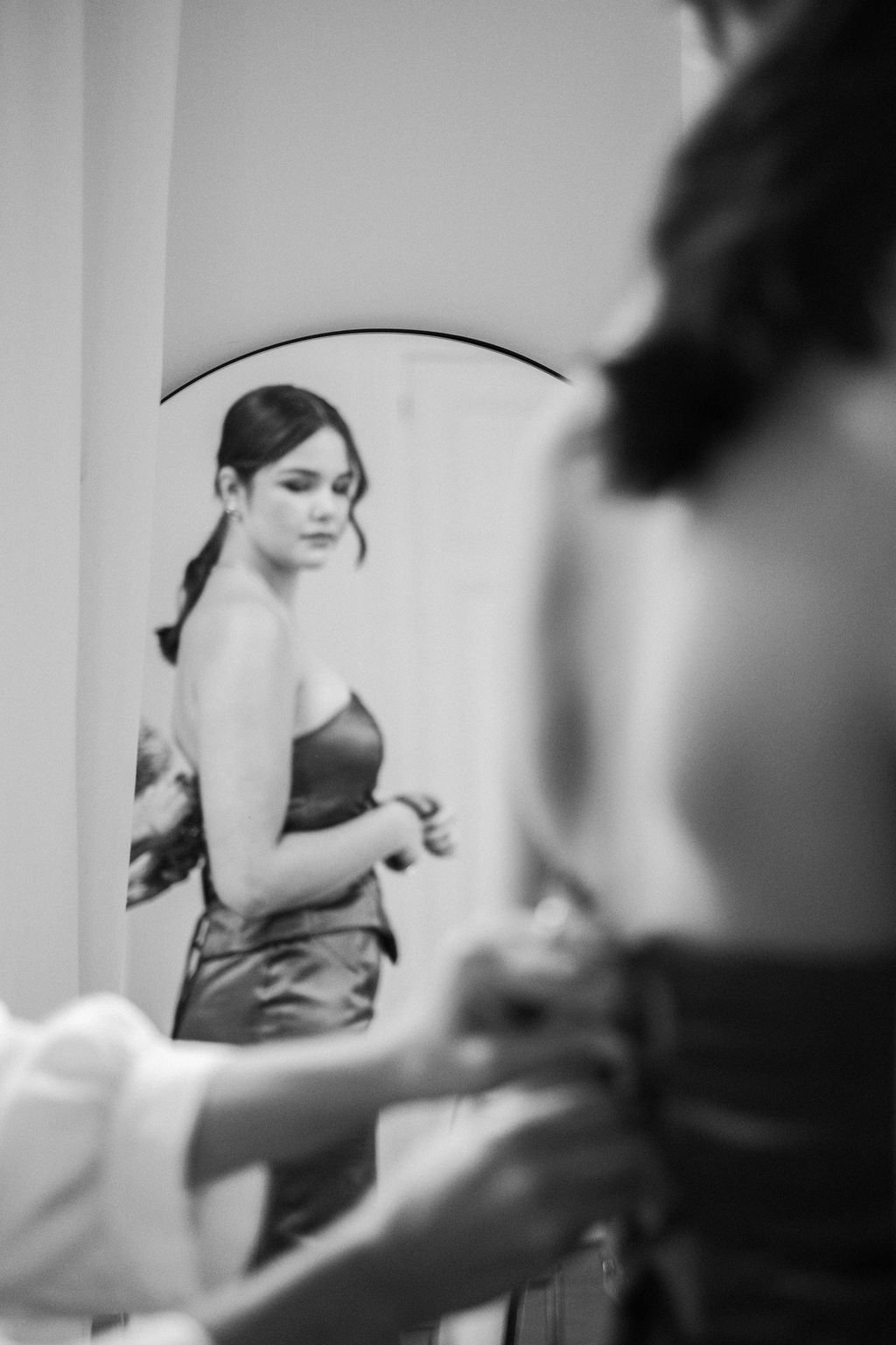 Black and white photo of a woman looking at herself in a mirror, with a blurred person in the foreground.