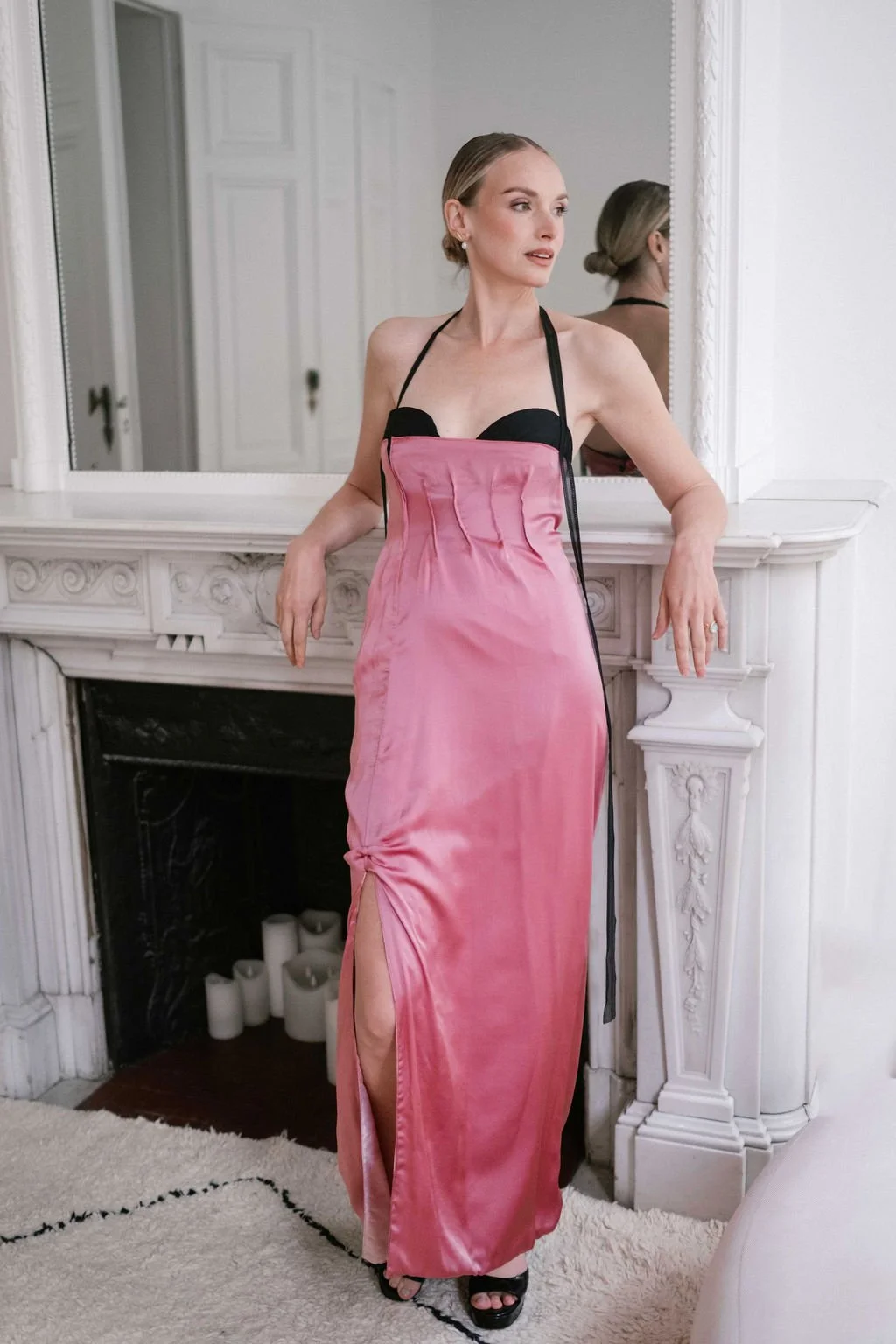 A woman in a pink satin dress with black straps posing in front of a mirror on a white fireplace mantel.