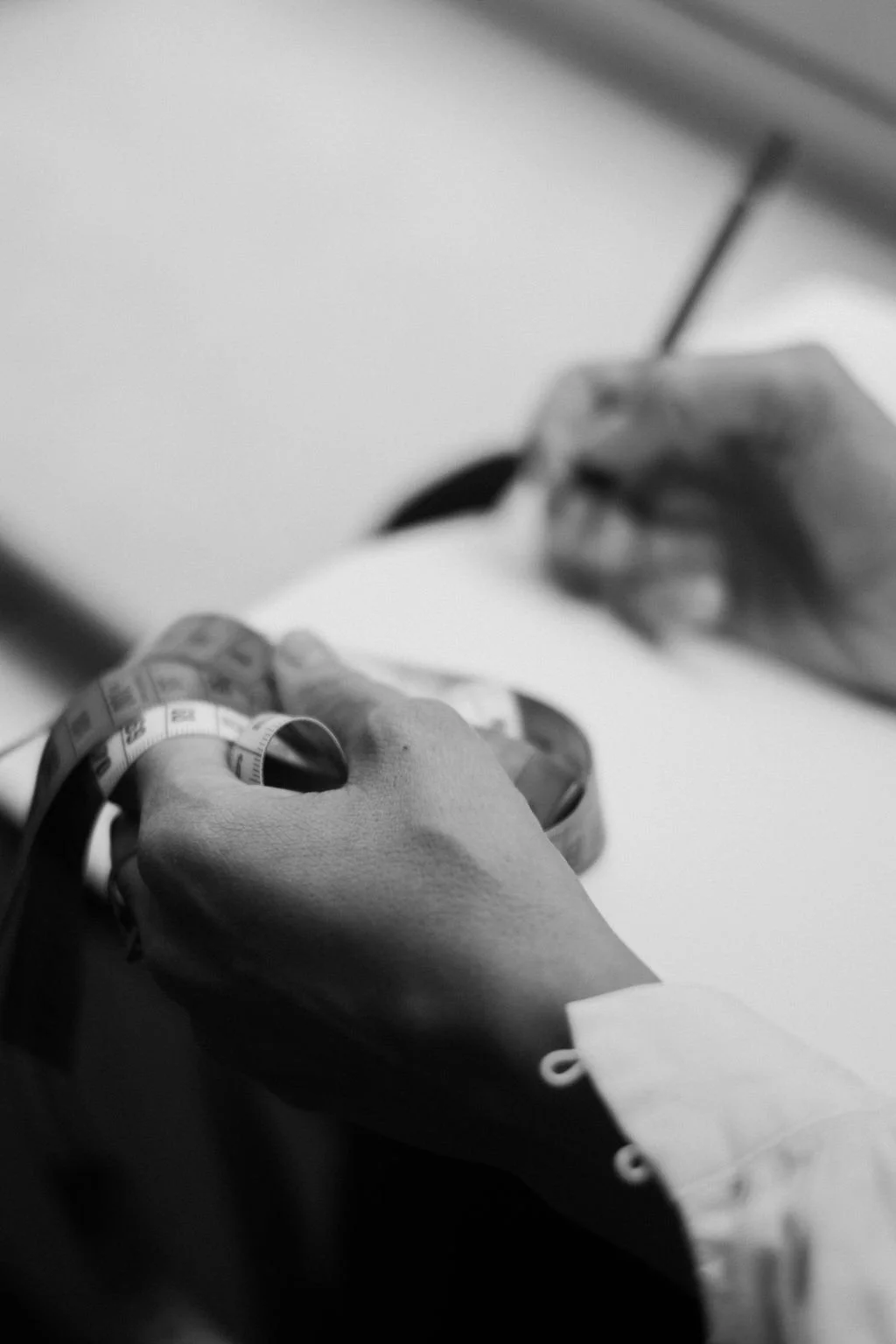 A close-up black and white photo of a person’s hands, one holding a measuring tape and the other writing with a pen on paper.