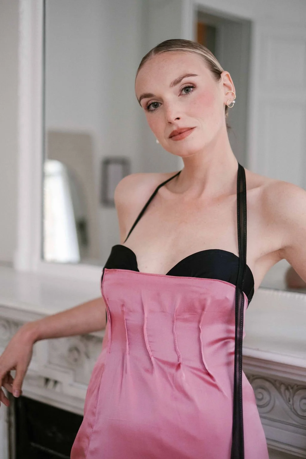 A woman with blonde hair and pearl earrings wearing a black and pink satin dress with spaghetti straps, standing in front of a mirror.