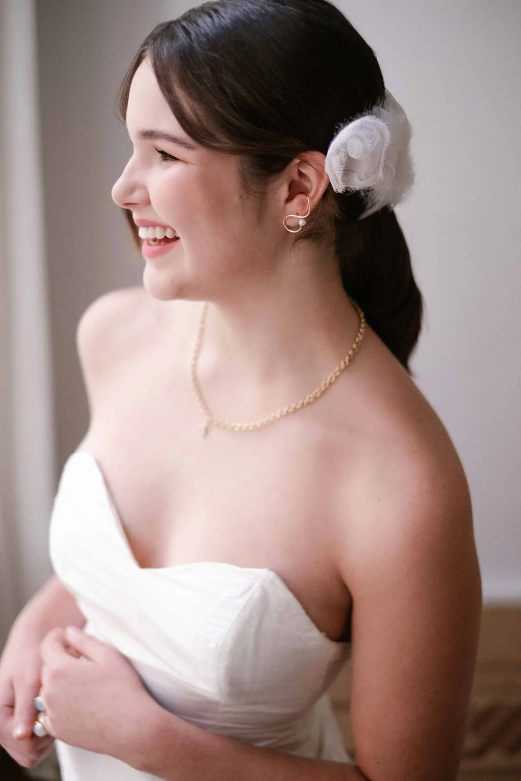A woman in a white strapless dress smiling, with gold jewelry and a hair accessory, looking to the side.