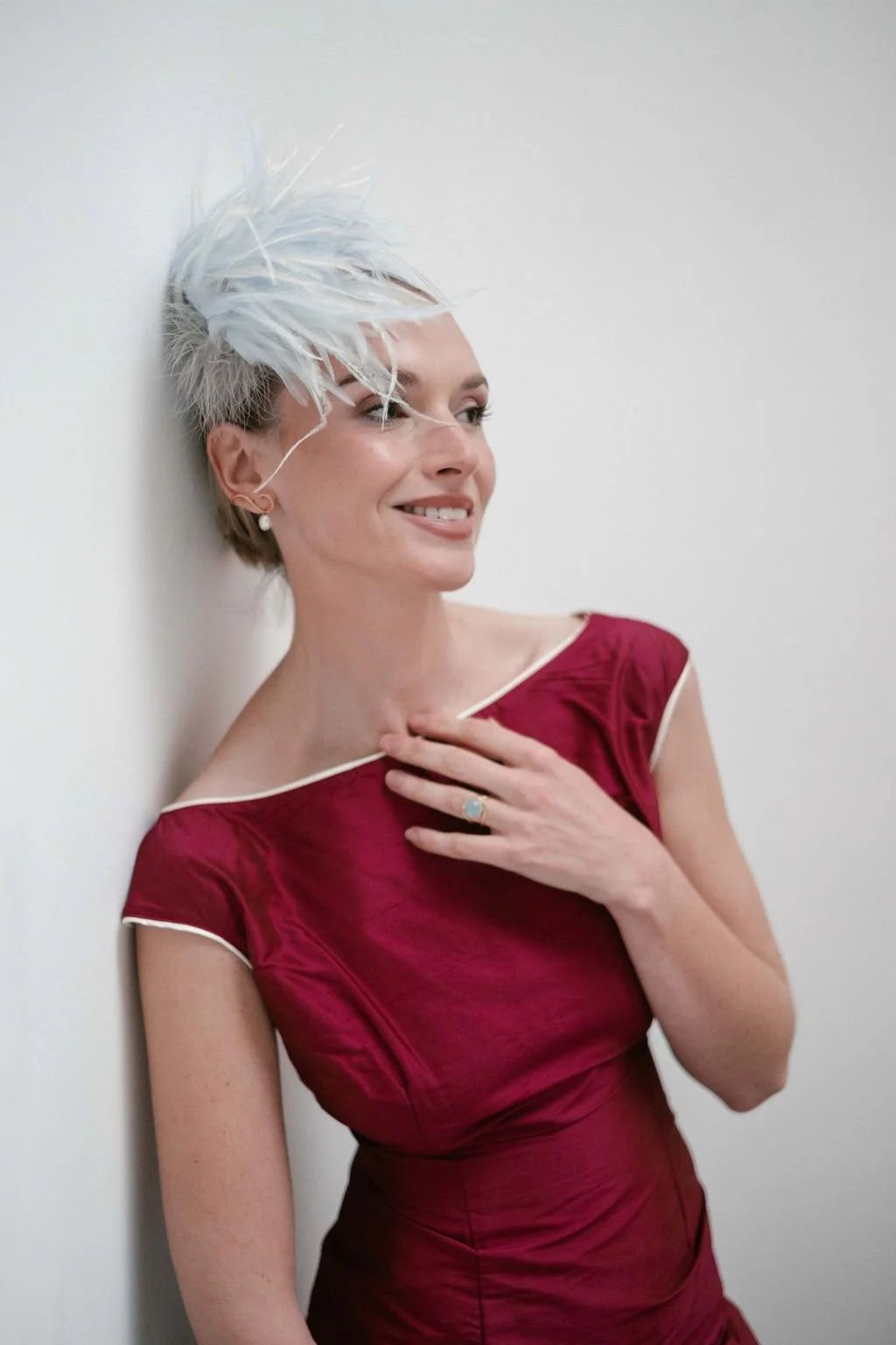 A woman with short, platinum blond hair styled upwards, smiling and leaning against a white wall, wearing a red dress with white piping and a ring with a large stone.