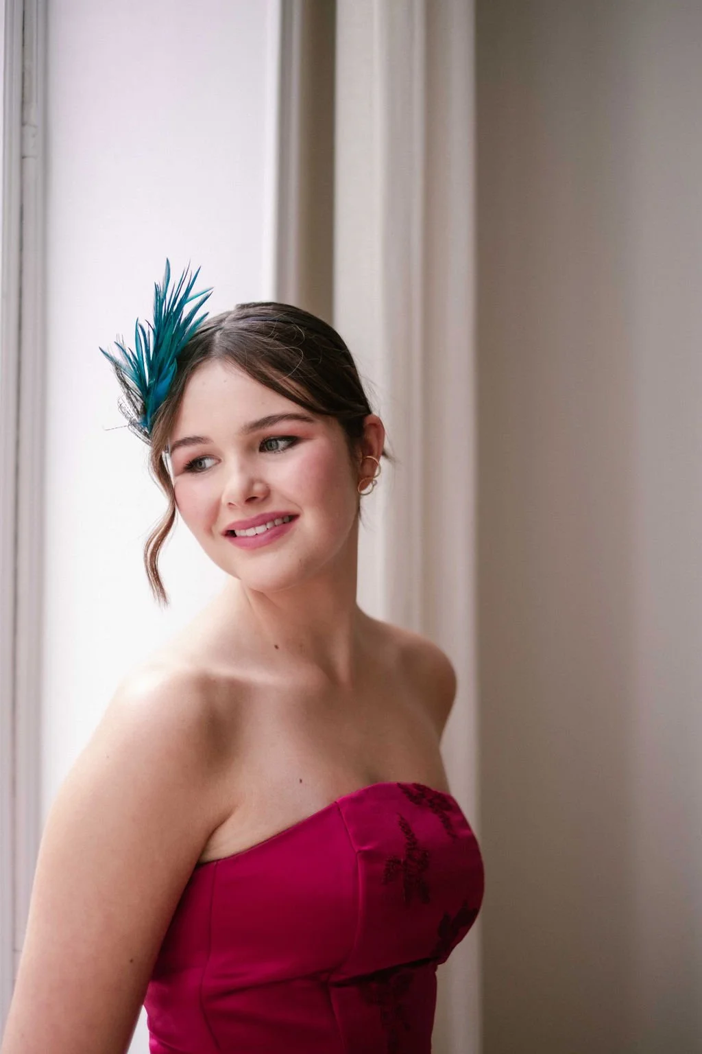 A young woman with brown hair styled in waves, wearing a strapless pink dress with black embroidery, a blue feather fascinator, and earrings, standing near a window with a neutral expression.