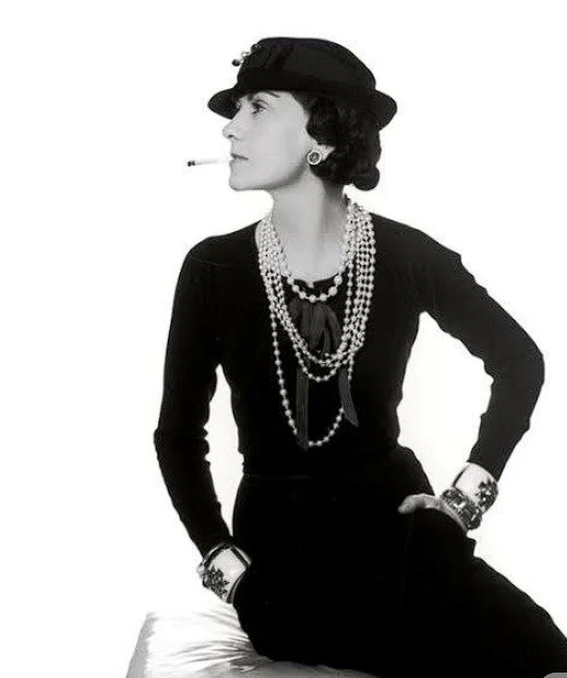 A woman sitting on a surface, wearing a black hat, black dress, and multiple pearl necklaces, with a cigarette in her mouth and jewelry on her wrists, posing in a vintage style.