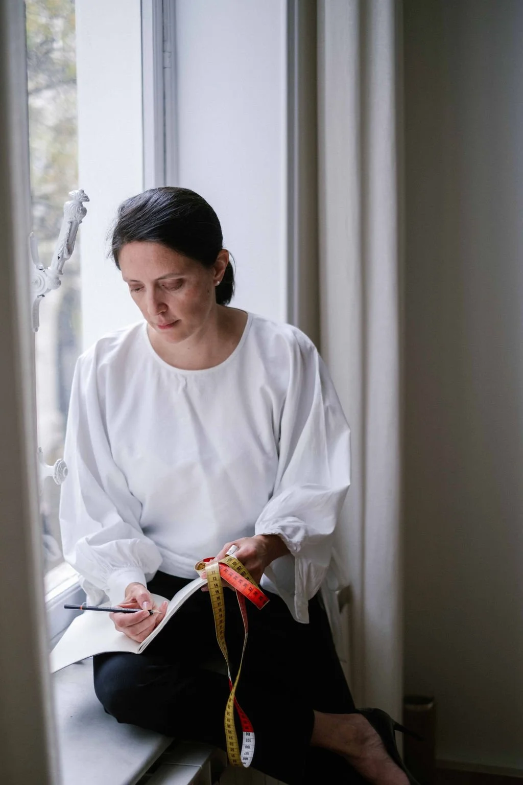A woman with dark hair sitting by a window, holding a tape measure and a pen, writing in a notebook.