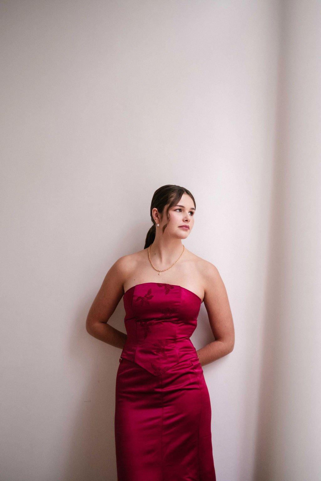 A woman in a strapless, red satin gown with embroidered floral details, standing against a plain off-white wall, looking to her right.