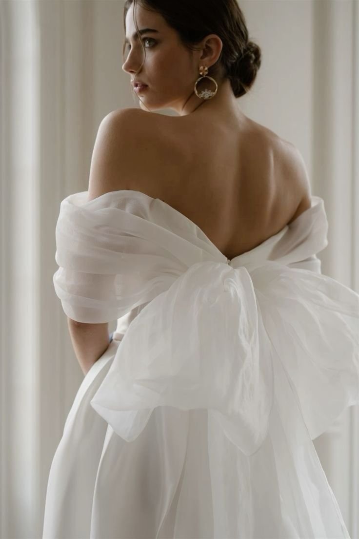 A woman with dark hair styled in an updo wearing a white off-the-shoulder dress with a large bow at the back, and gold hoop earrings, standing near a window.