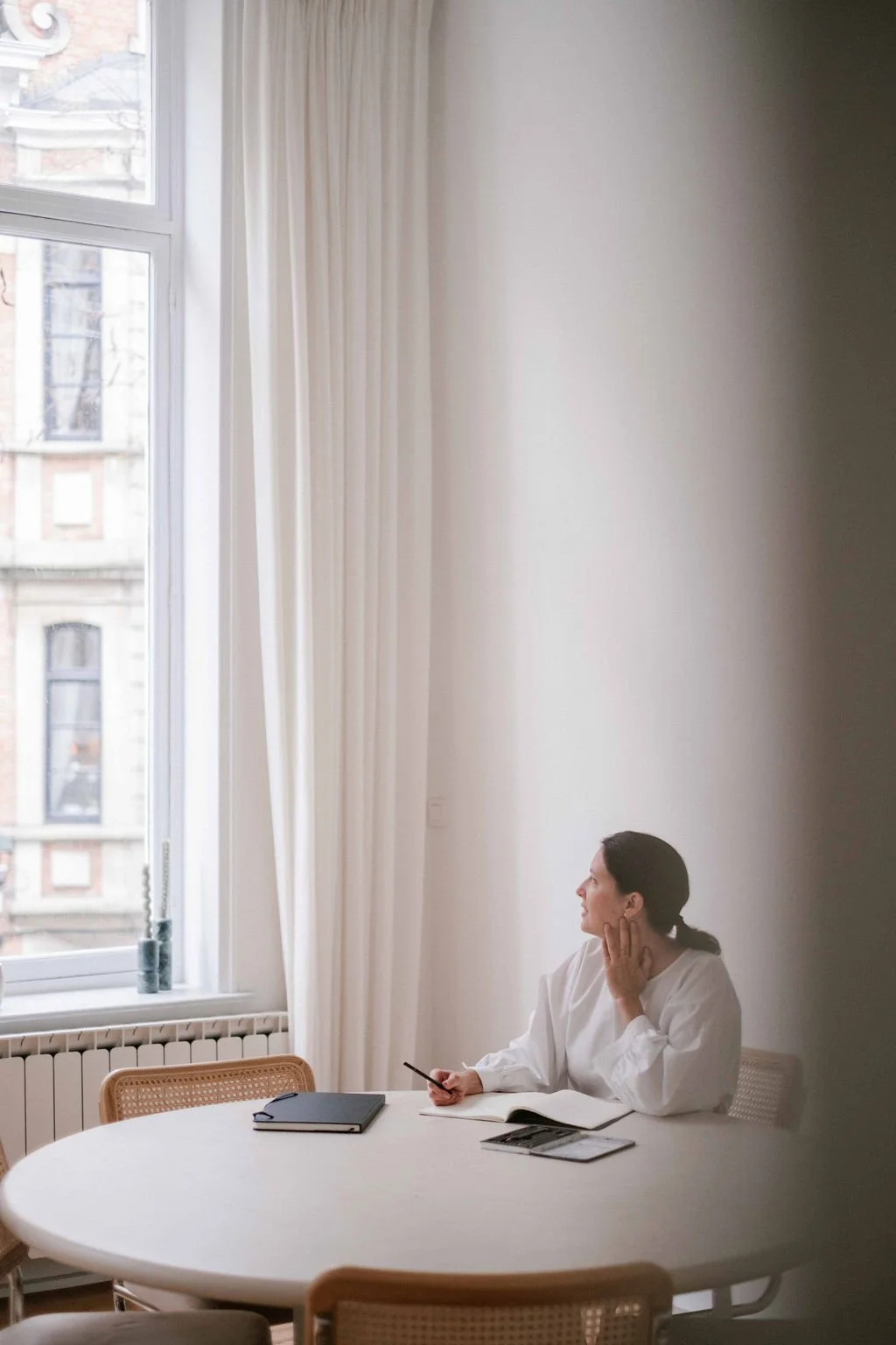 A woman sitting at a round table near a large window, holding a pen and looking to the side, with an open notebook, a closed notebook, and a magazine on the table.