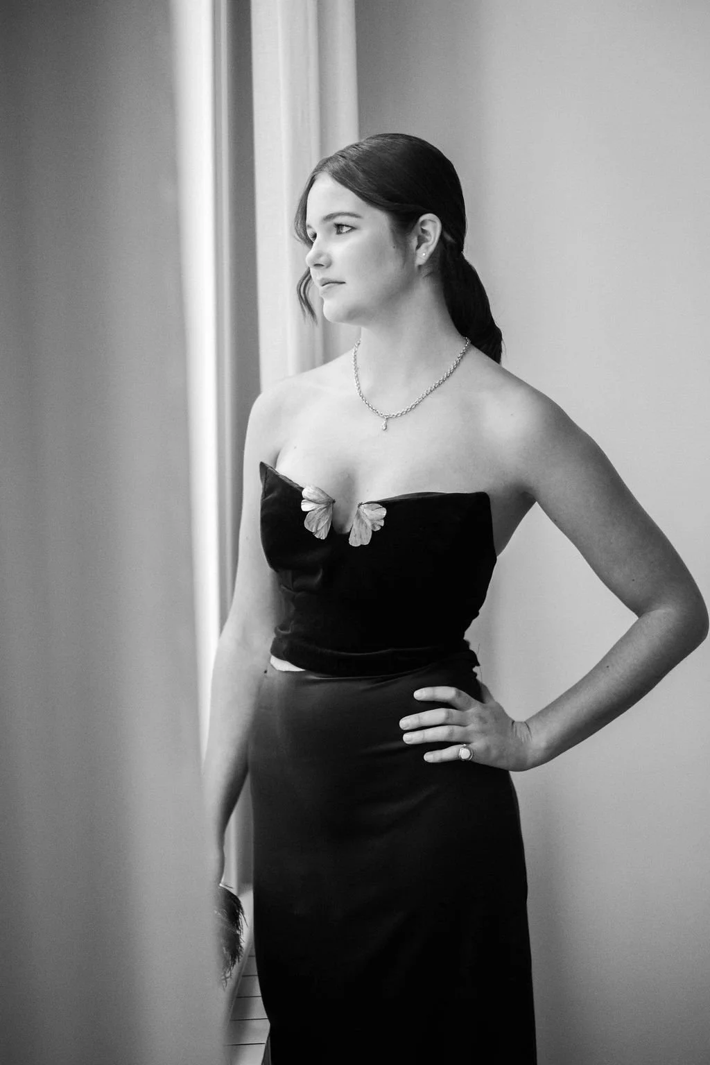 A woman in a strapless black dress with butterfly embellishments at the top, standing in profile near a window, looking pensively outside, with her right hand on her hip.