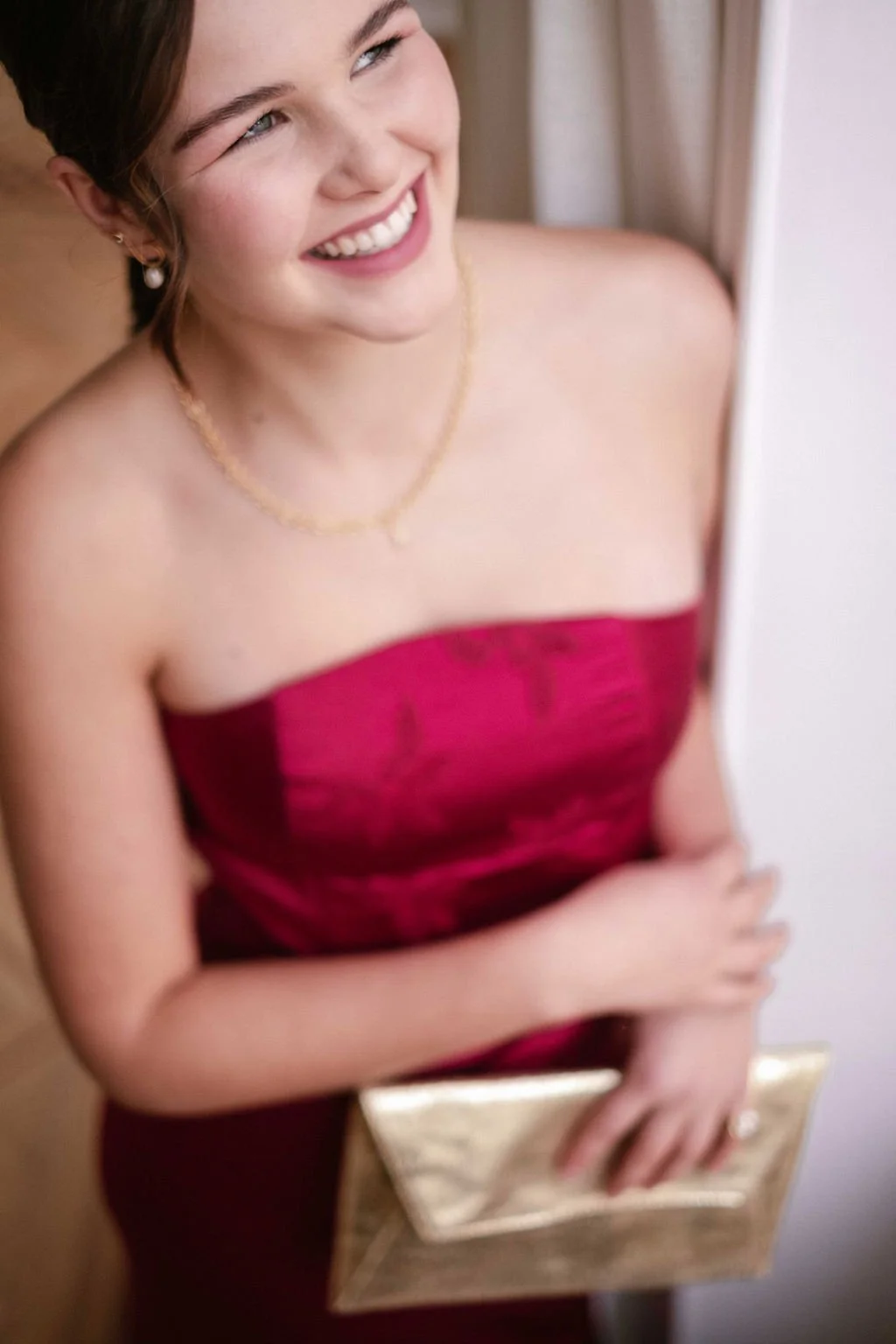 A young woman with dark hair and fair skin, wearing a strapless red dress, smiling, holding a gold clutch, standing indoors near a white wall.