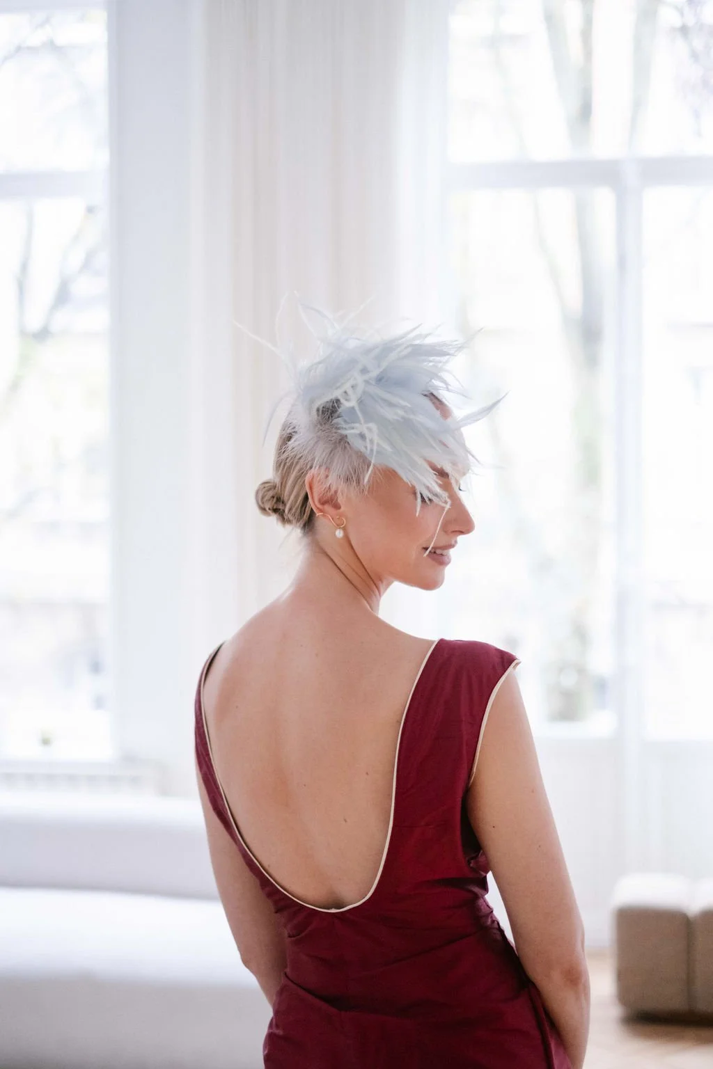 A woman in a sleeveless, deep red dress with a low back, wearing pearl earrings and a large white feathered headpiece, stands indoors near white curtains and large windows with natural light.
