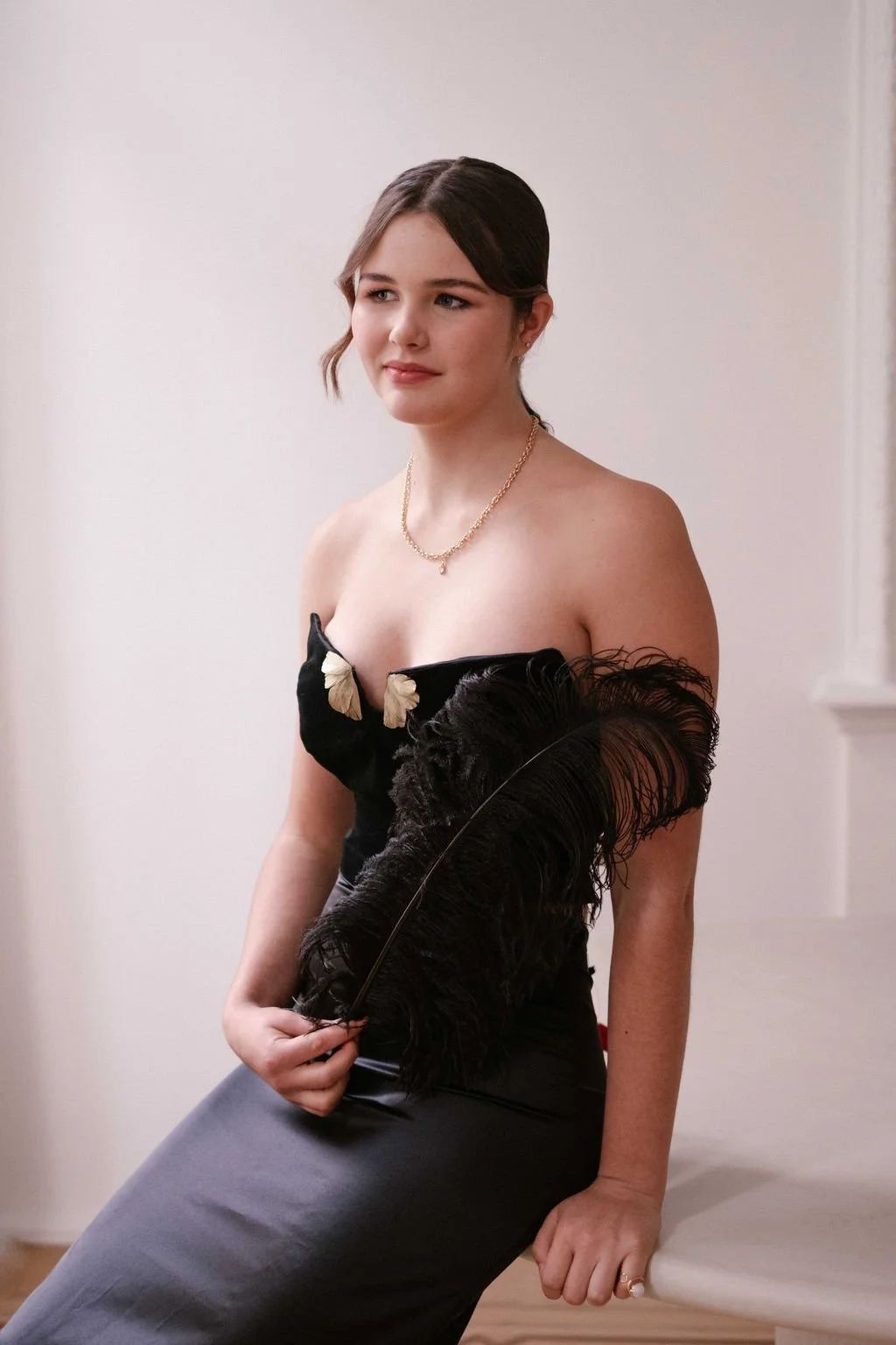 A young woman with brown hair, wearing a black strapless dress with butterfly details, holding a large black feathered fan, and sitting against a white wall.