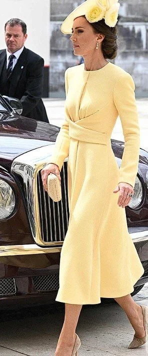 A woman in a yellow coat, hat with flowers, and heels walking next to a luxury car, with a man in a suit in the background.