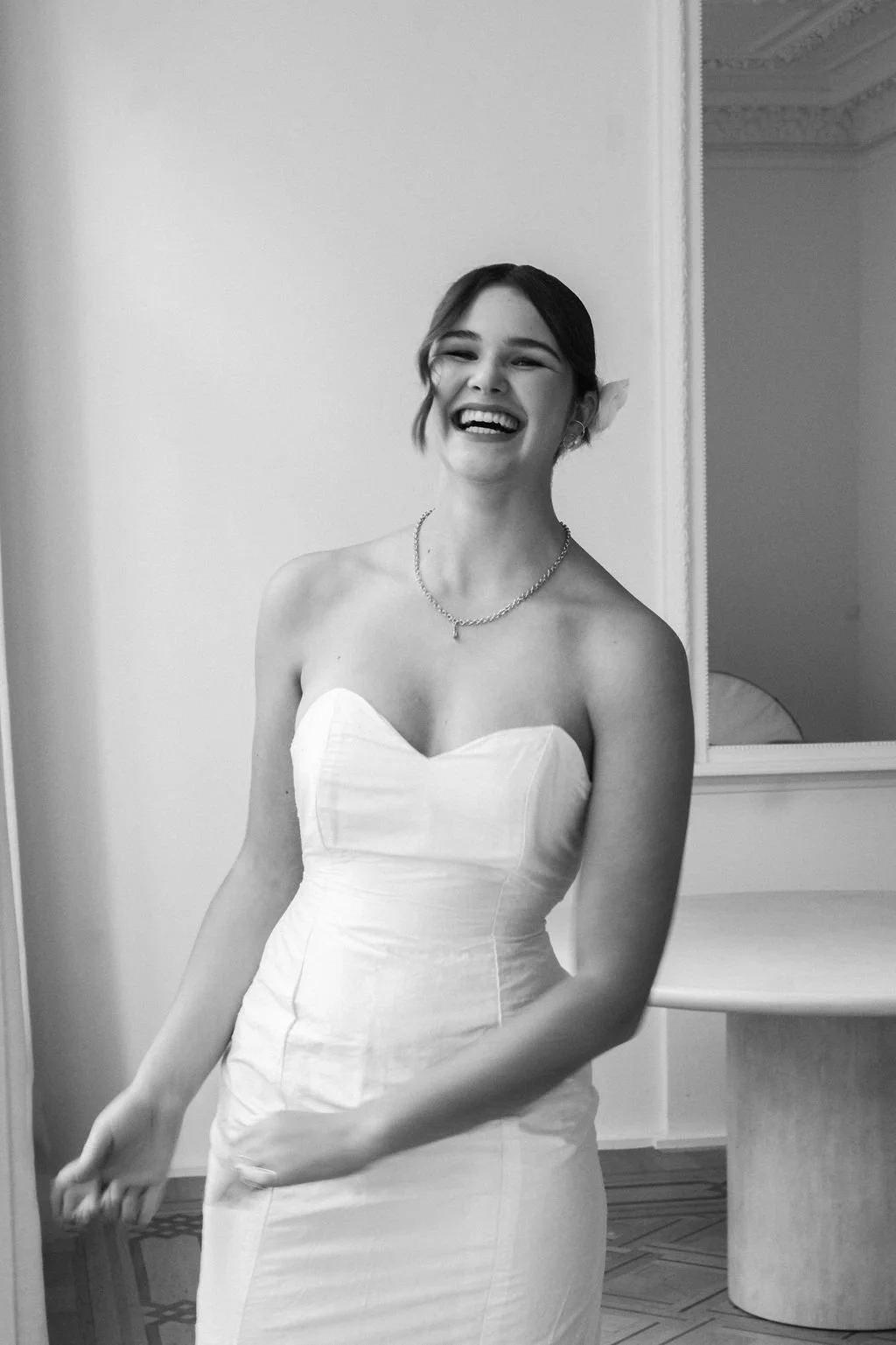 A smiling woman in a strapless white dress and necklace, standing indoors near a mirror.