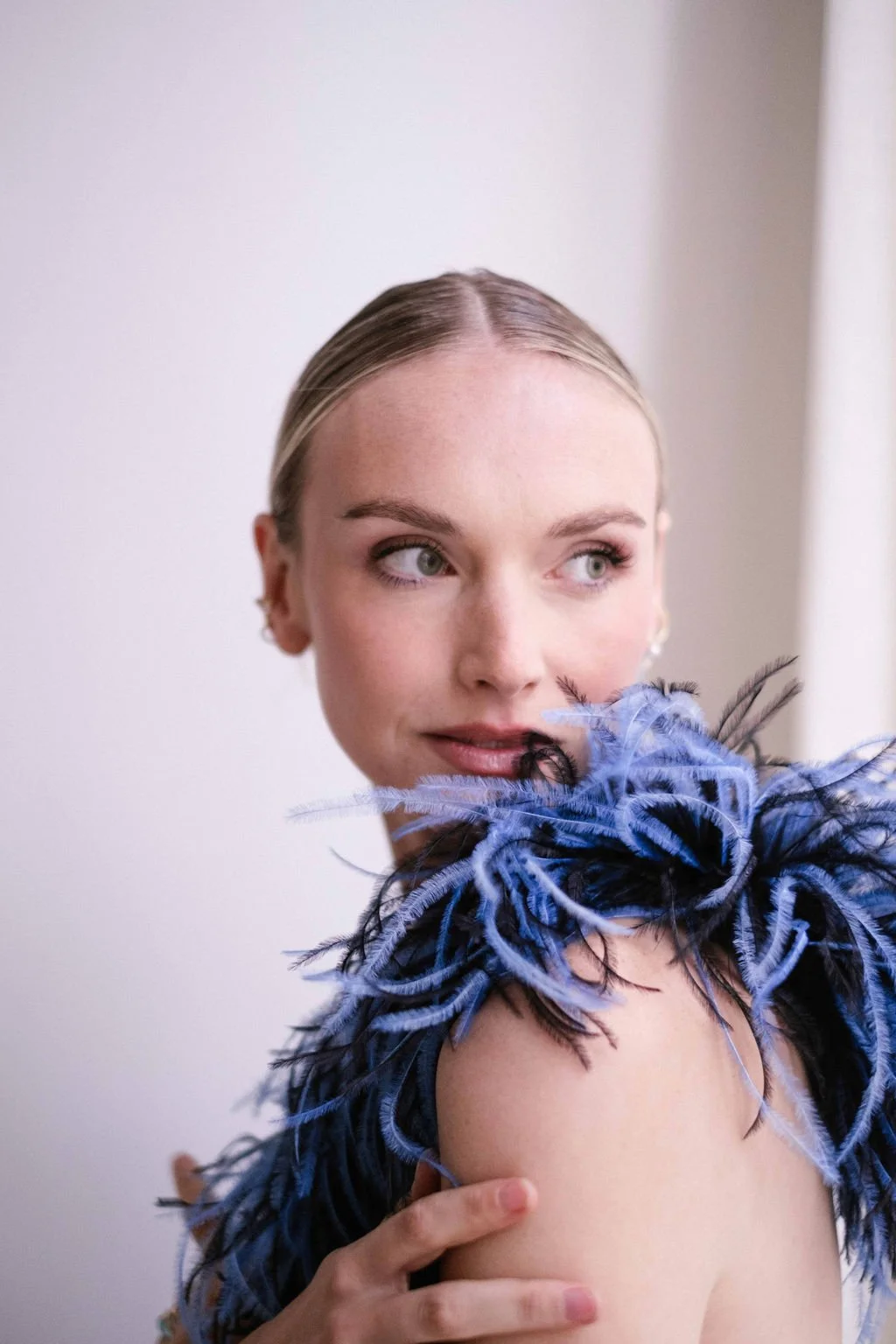 A woman with blonde hair and green eyes is holding a black and blue feathered accessory on her shoulder and looking to her left. The background is plain and light-colored.