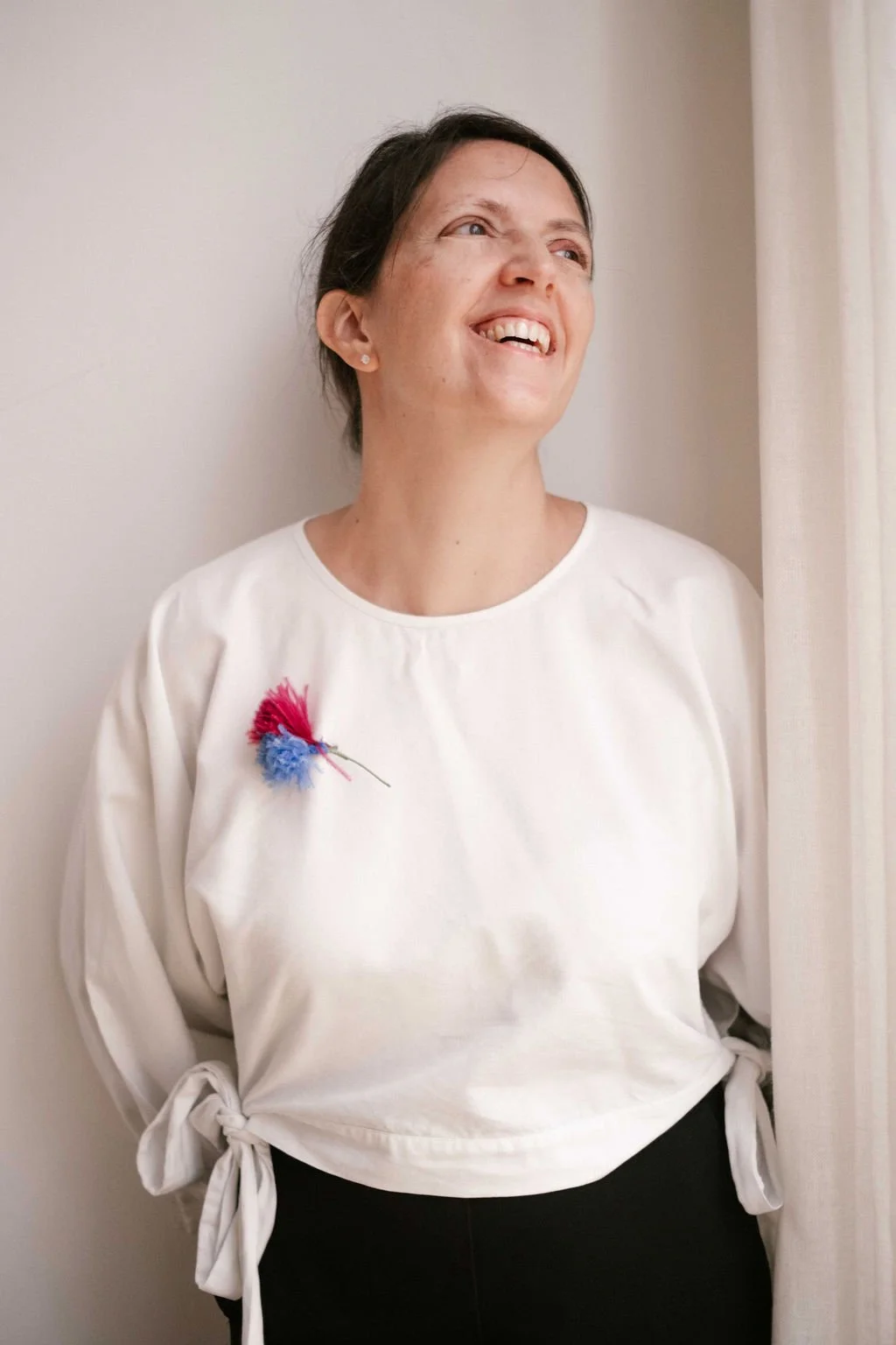 A woman with short dark hair and fair skin smiling and looking to her right, standing next to a white curtain, wearing a white top with a pink and blue pin in the shape of a flower on her left side.