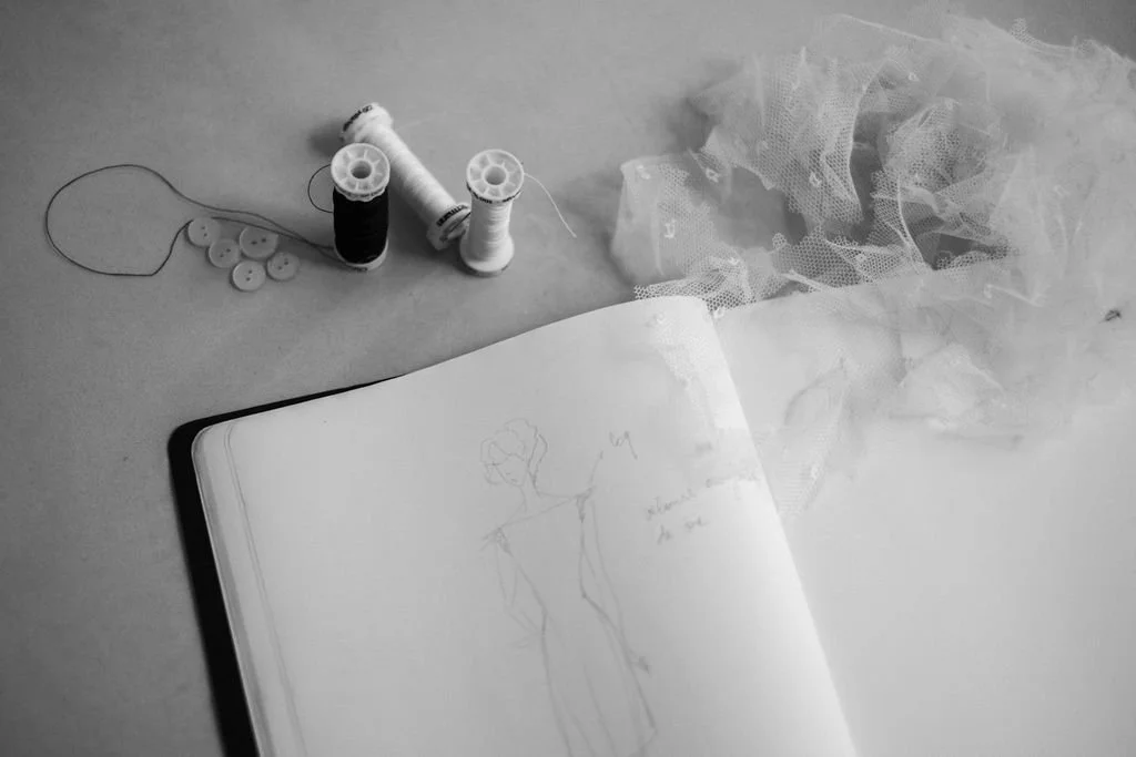 A sewing kit with spools of thread, buttons, a piece of tulle fabric, and a sketchbook with a fashion design drawing on a flat surface.