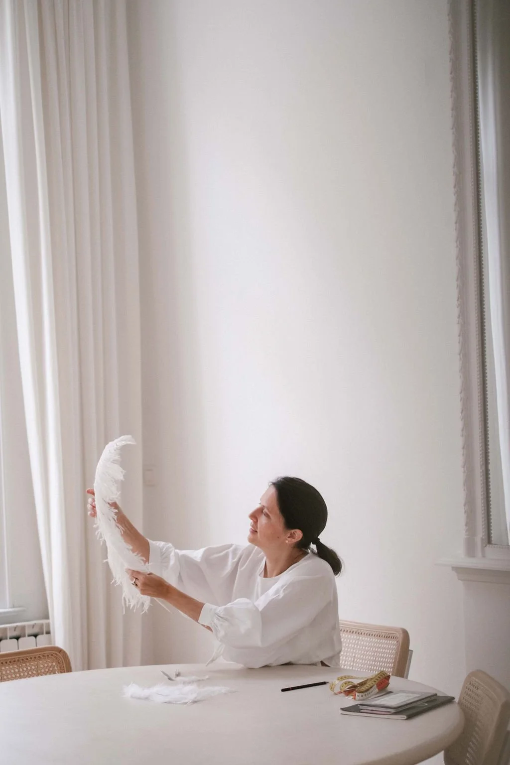 Woman sitting at a table in a bright room, holding a white feathered object, with a measuring tape, notebook, and pen on the table.