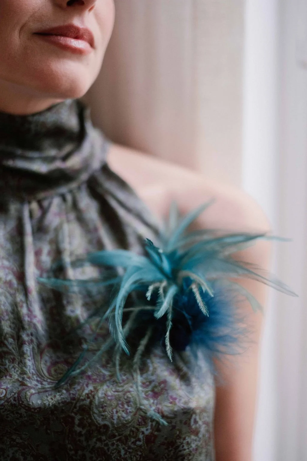 Close-up of a woman wearing a patterned dress with a feathered blue accessory on her shoulder, near a window.
