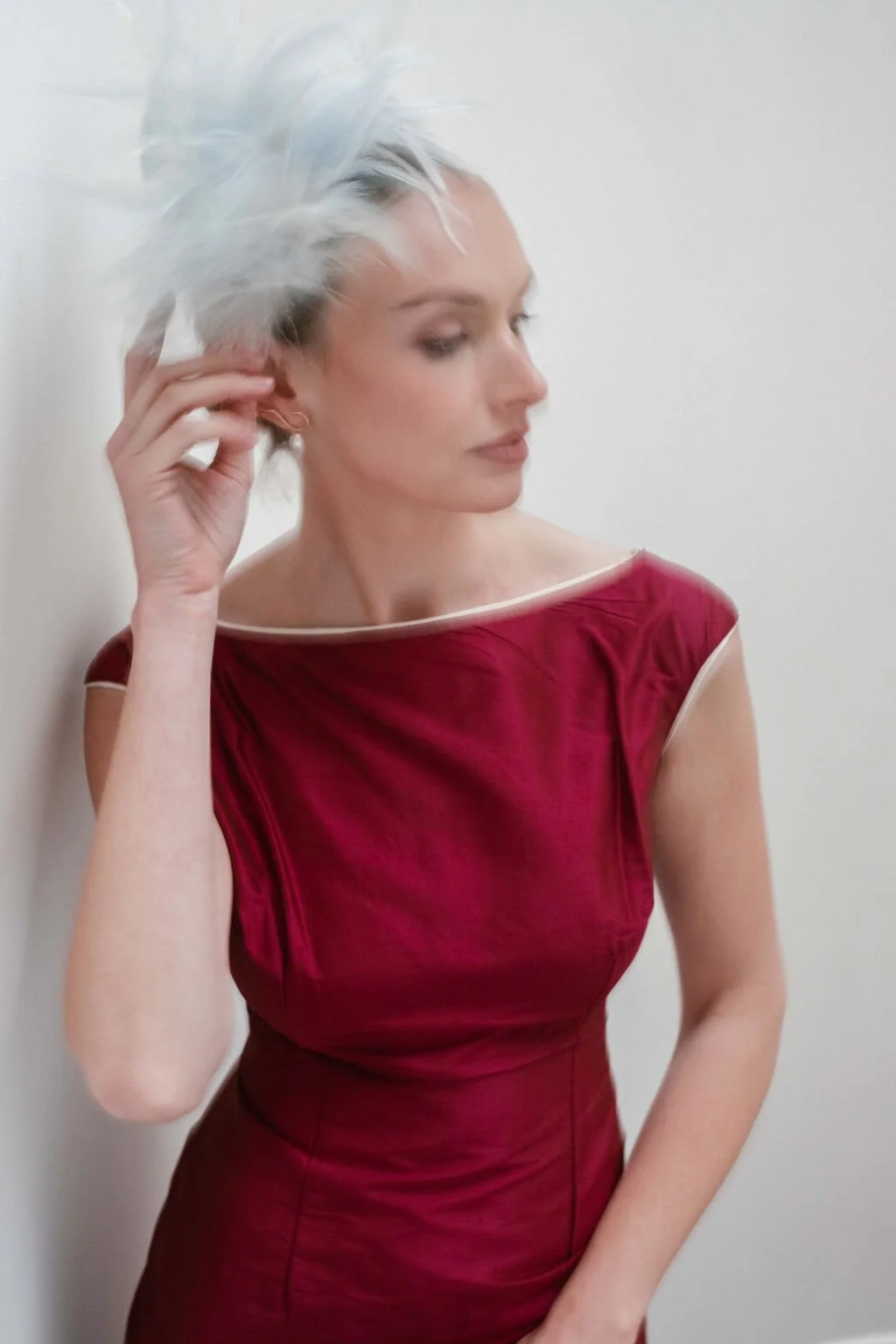 A woman in a red dress with white trim adjusting her white hair, standing against a plain white wall.