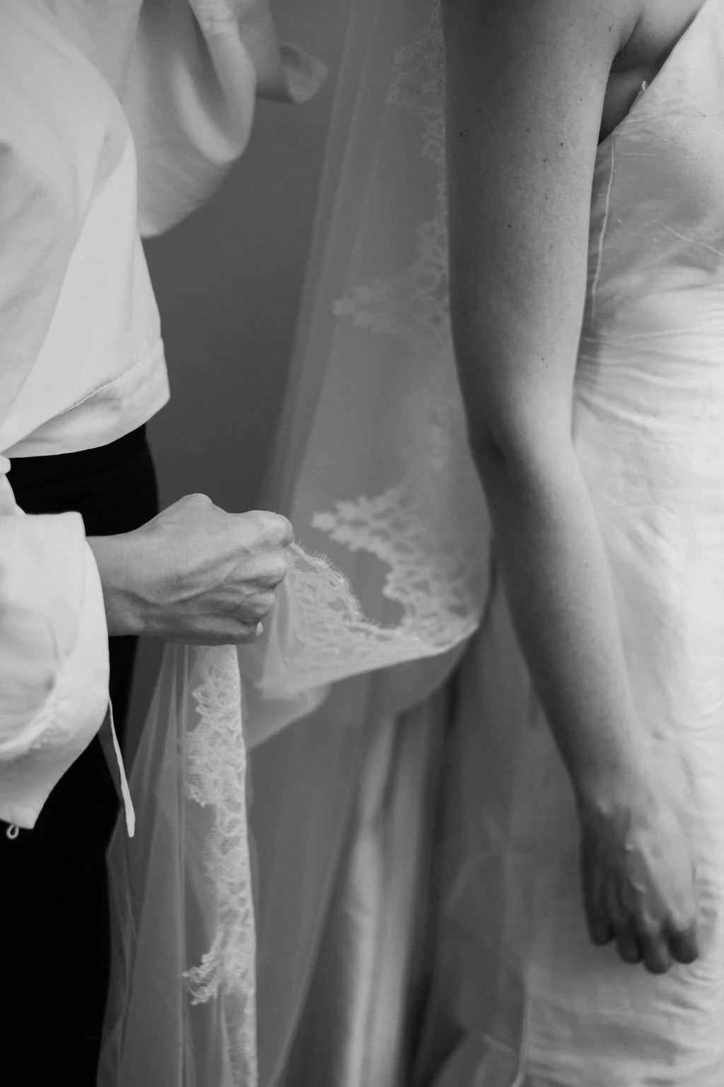 Close-up of a person holding lace fabric beside a bride wearing a wedding dress.