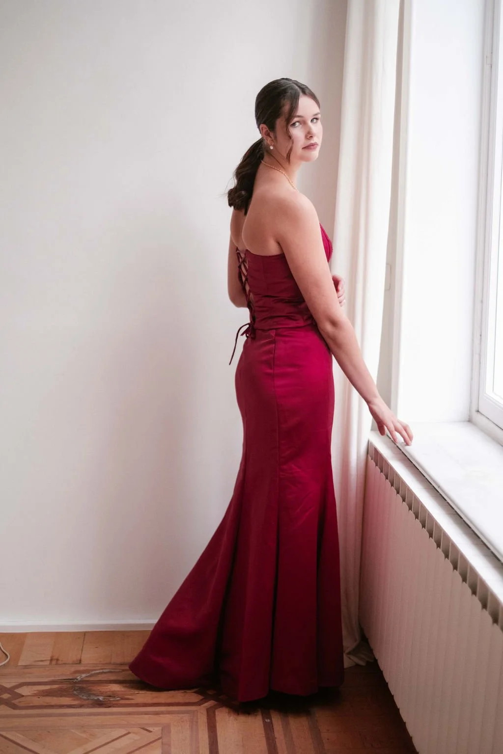 A woman in a strapless, form-fitting red gown standing by a window, looking at the camera with her hand resting on the window sill.