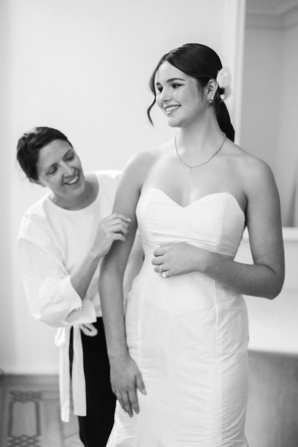 A woman in a strapless wedding dress smiling as she gets dressed and assisted by another woman who is helping her with the gown.
