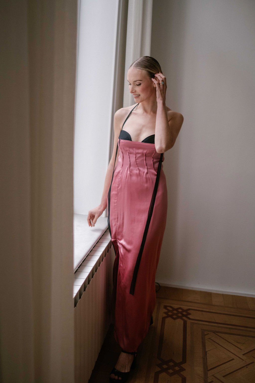A woman in a pink satin dress with black accents, standing near a mirror and adjusting her hair.
