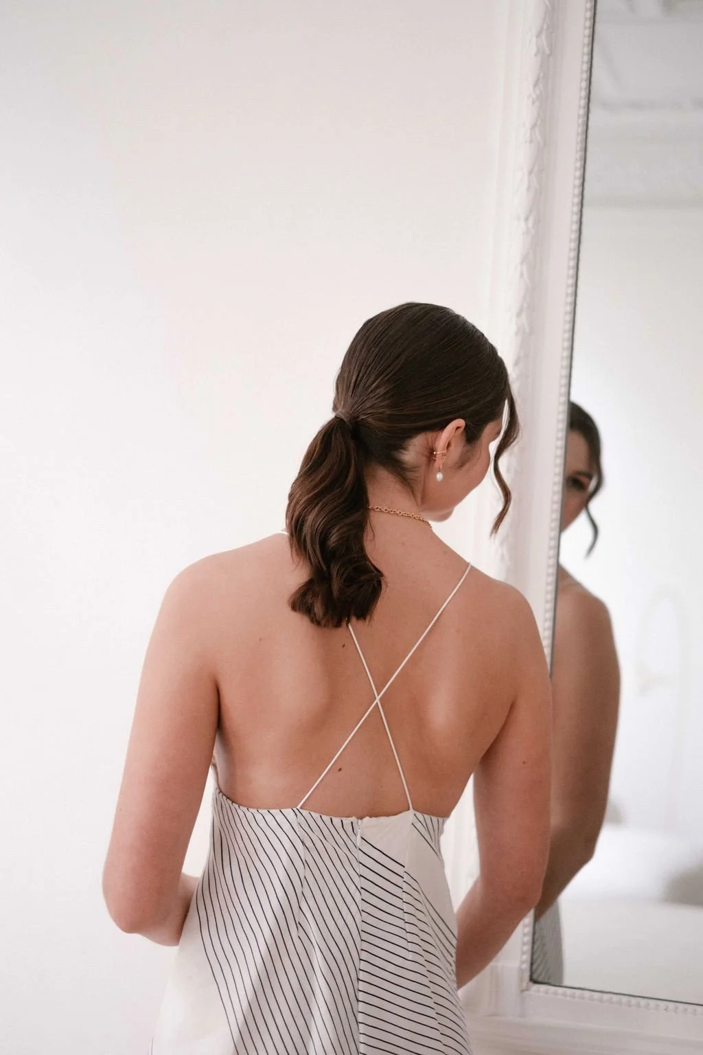 Back of woman with wavy brown hair in a low ponytail, wearing a white dress with thin spaghetti straps, standing in front of a mirror.
