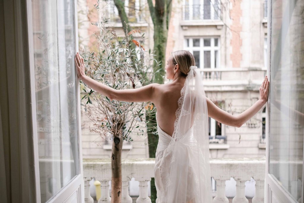 A bride in a white wedding dress and veil standing on a balcony with her arms outstretched, looking outside at buildings and trees.