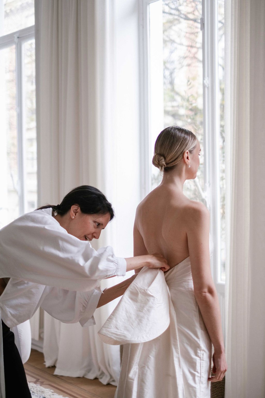 A woman in a strapless wedding dress is assisted by a stylist as she gets ready, with large windows and white curtains in the background.