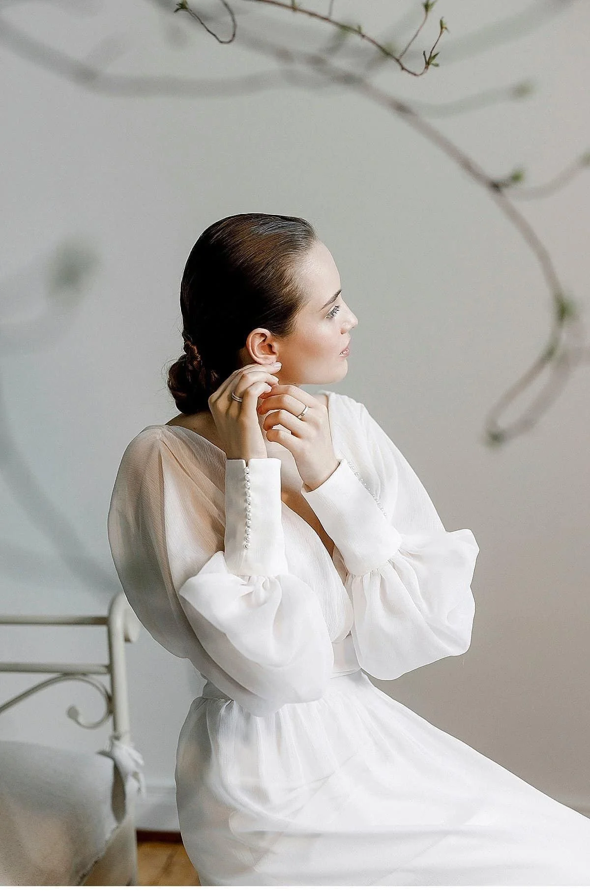 A woman with brown hair styled in a braid sits in a white dress, looking to her right, touching her ear with her left hand, indoors with a simple background.