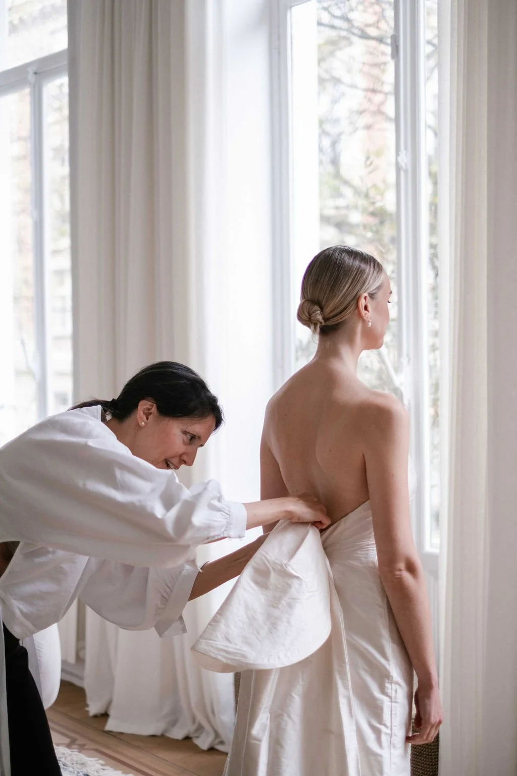 A woman in a wedding dress is having her gown adjusted by a stylist in a bright room with large windows and white curtains.