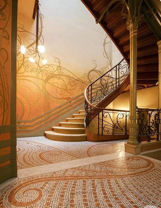 Elegant spiral staircase with ornate black railing in a warmly lit lobby with patterned tile flooring and intricate gold-colored wall art.