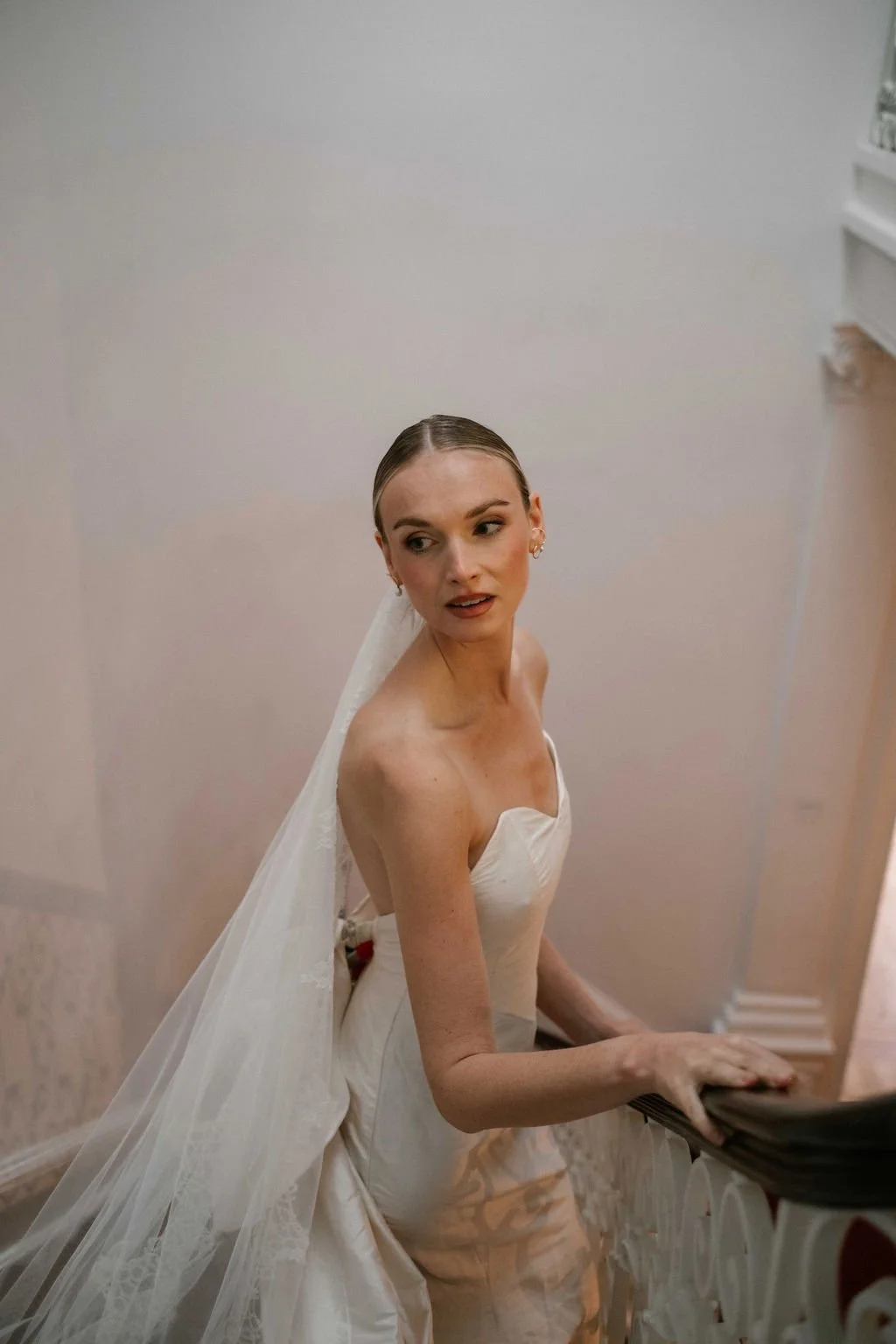 A woman in a white wedding dress with a veil sitting on a staircase, looking off to the side.