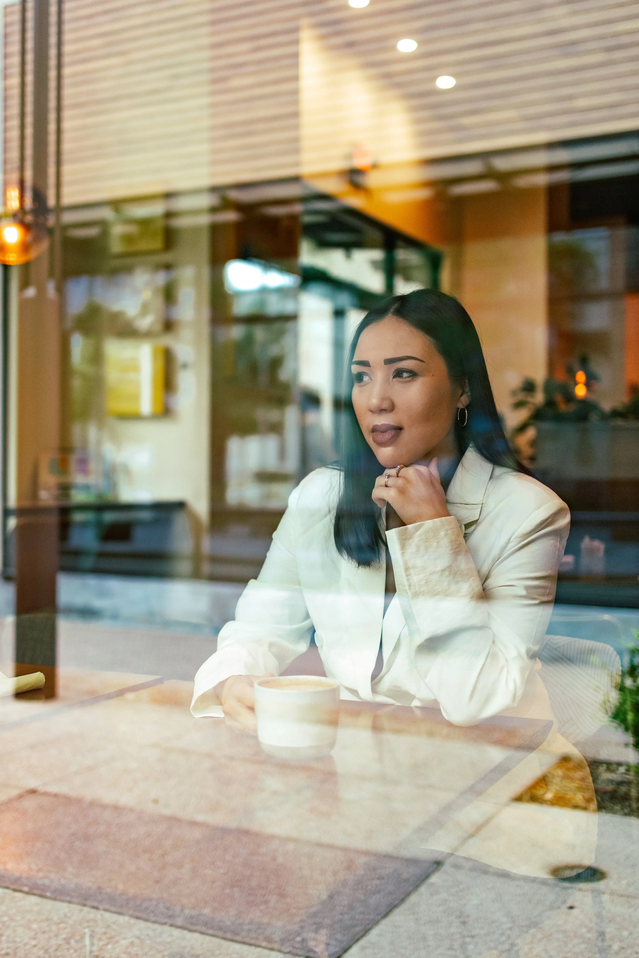 A therapist in Texas sitting inside a cafe, looking out through a window with a cup of coffee in front of her on the table.