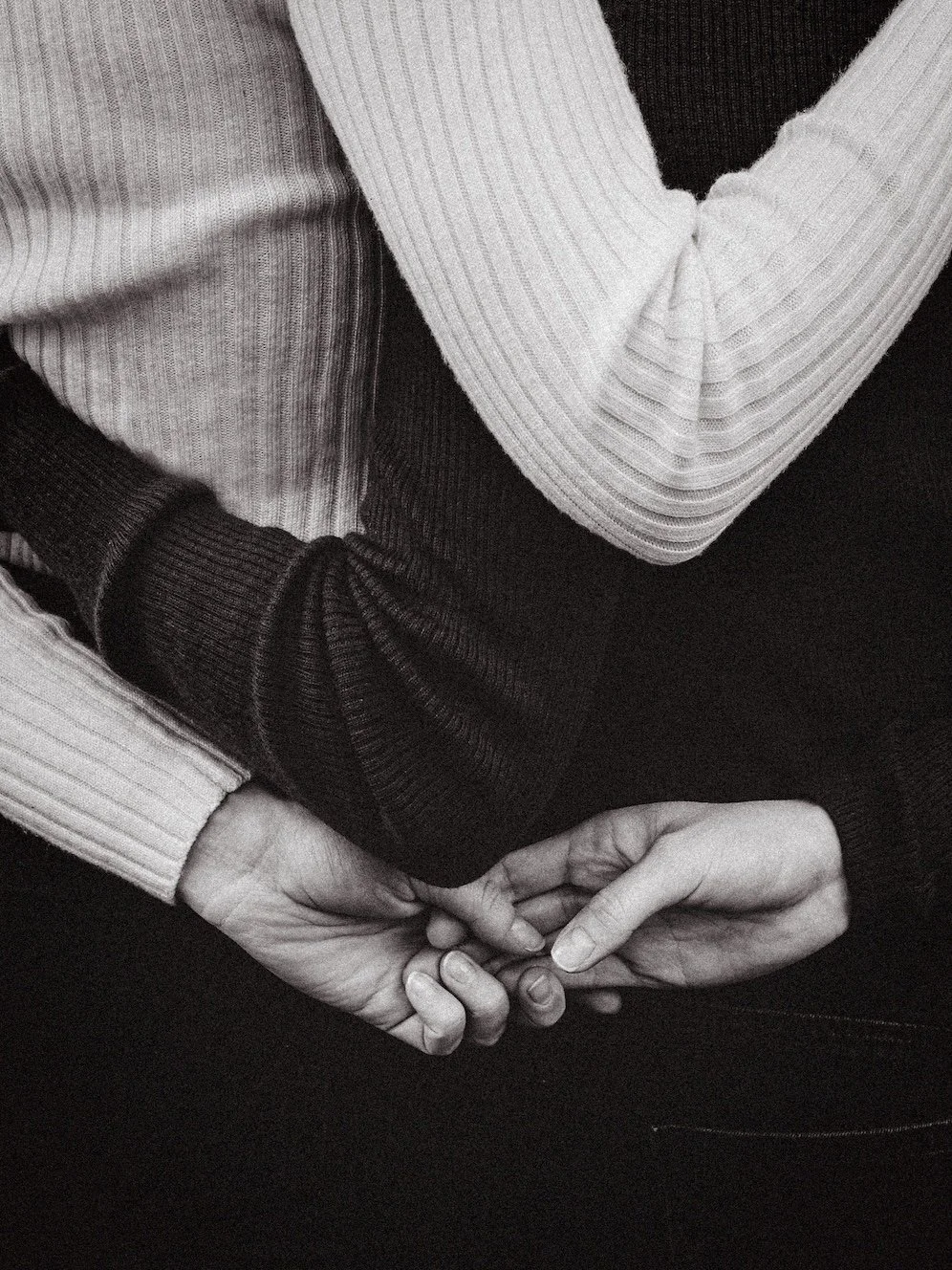 Close-up of two people holding hands, with one person's hand resting on the other's wrist, in black and white.