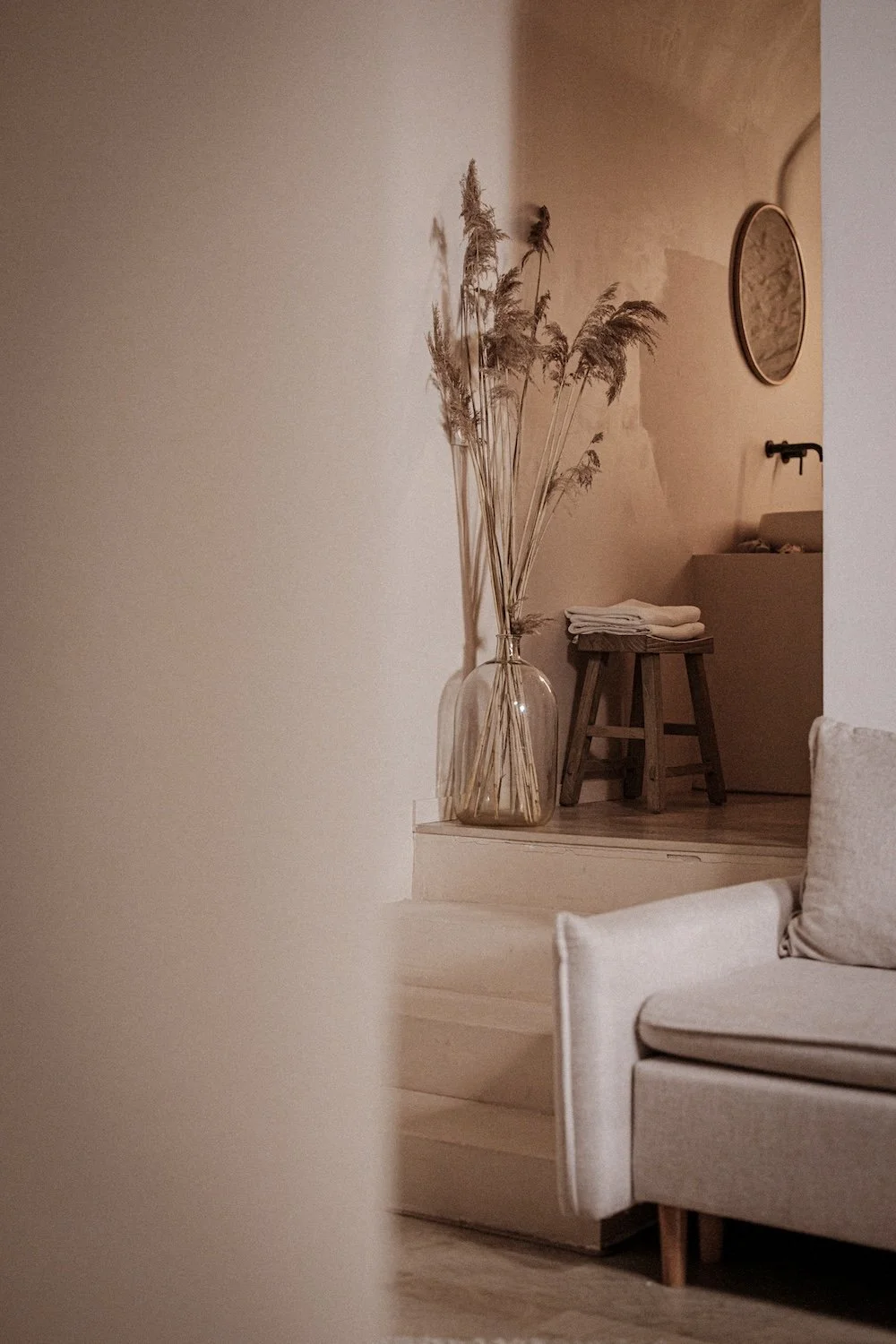 A cozy, minimalist living room corner with a beige sofa and a glass vase of dried pampas grass on a small wooden platform.