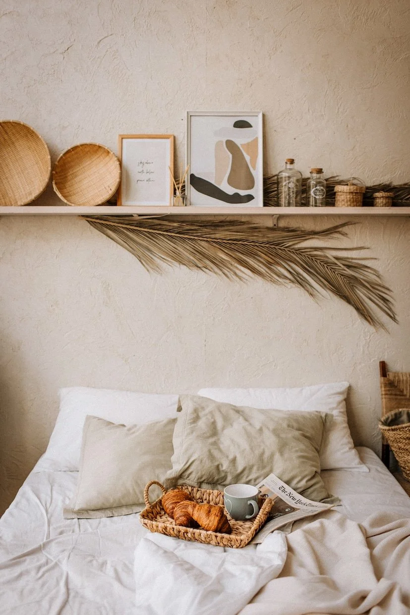 A cozy bed with beige pillows and a white bedspread, a woven tray with a croissant, a mug, and a newspaper on it, above the bed a floating shelf holds framed art, woven baskets, glass jars, and a large dried palm leaf across the wall.
