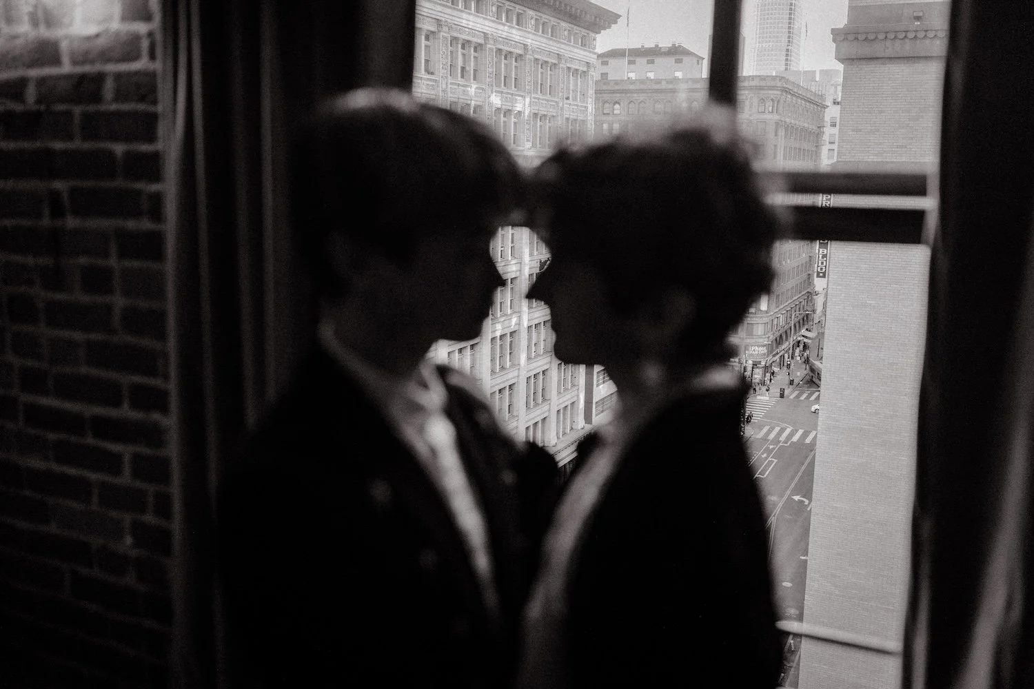 Silhouettes of two people close to each other in front of a window, with an urban cityscape visible outside, in black and white.