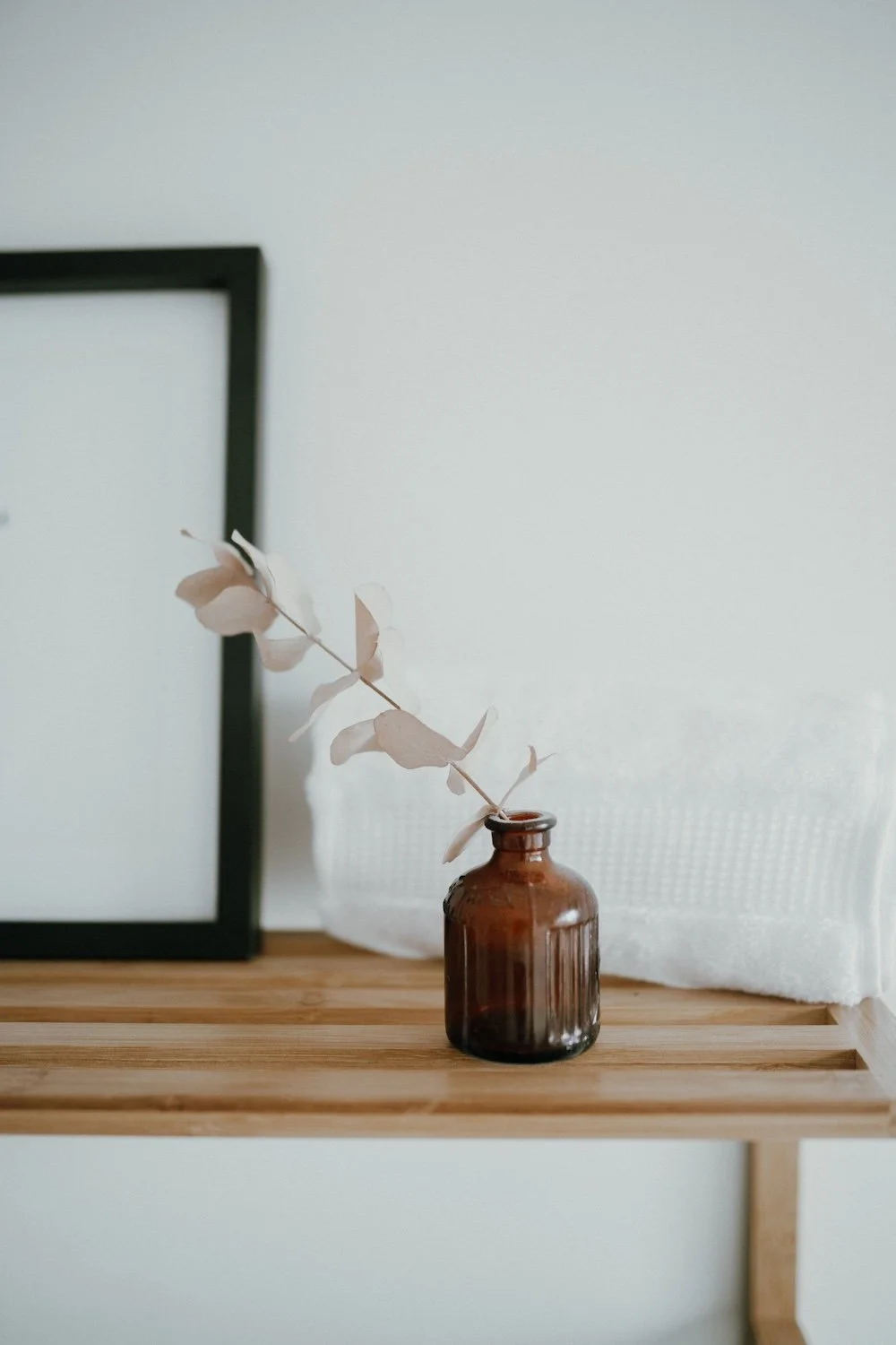 A brown glass vase with a dried eucalyptus branch on a wooden surface against a white wall.