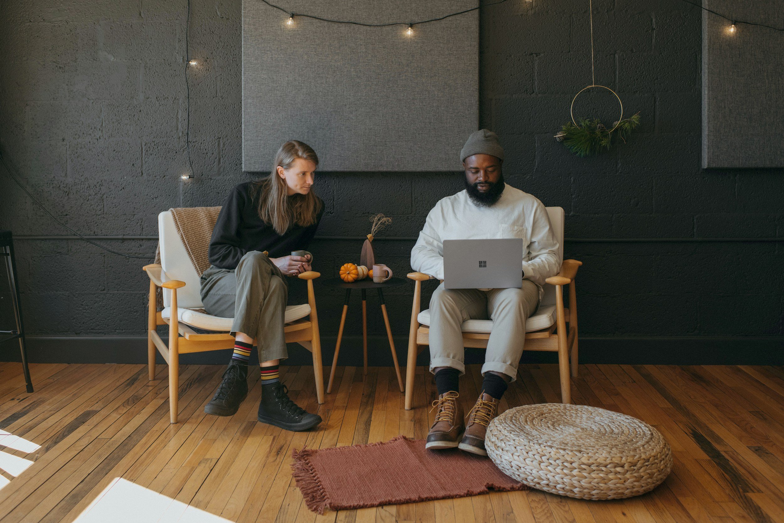 Two people seated on chairs in a modern room; one person is a woman with long hair in a black top and khaki pants holding a cup, and the other is a man with a beard in a white shirt, gray beanie, and tan pants using a laptop; small side table with pumpkins and decor between them, ambient string lights overhead, and a woven floor cushion on the wooden floor.