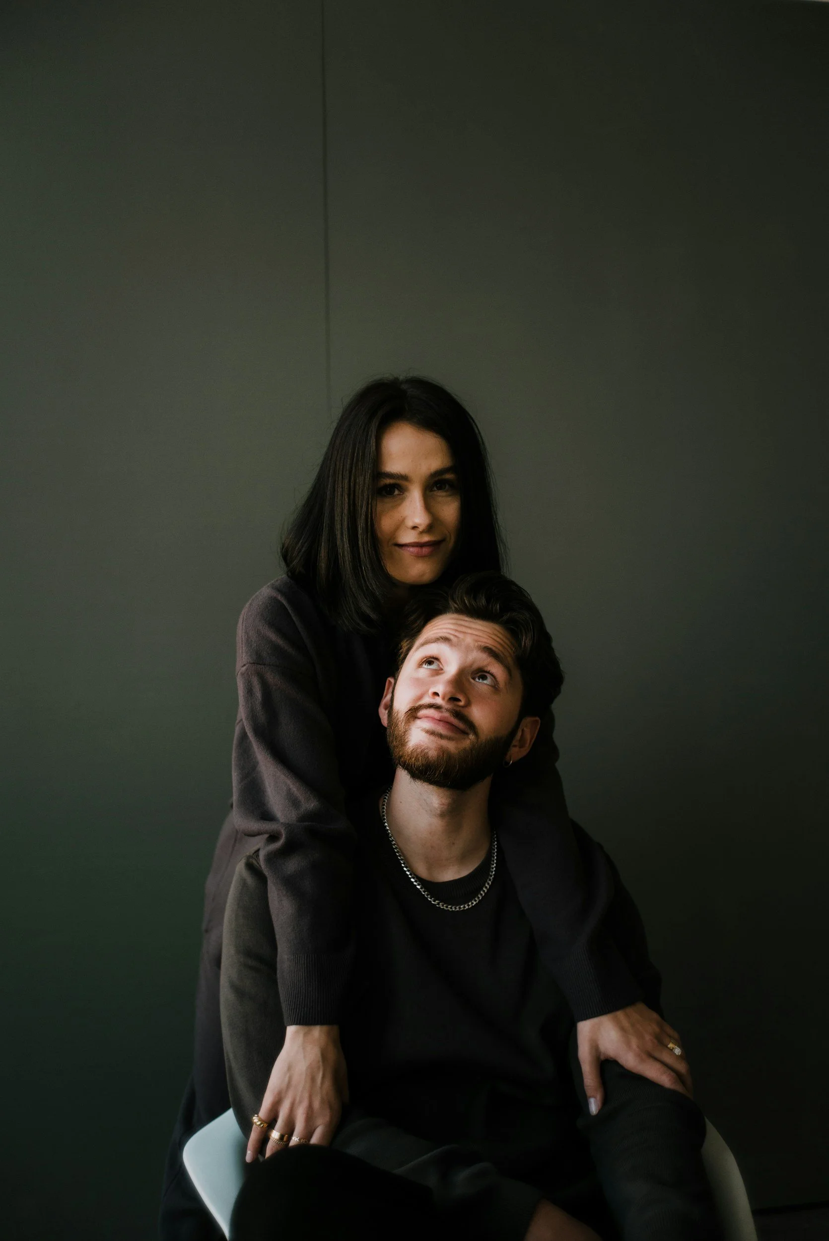 A woman standing behind a man, both dressed in dark clothing, against a dark background. The woman has short black hair and the man has a beard and mustache, looking up at her while smiling.
