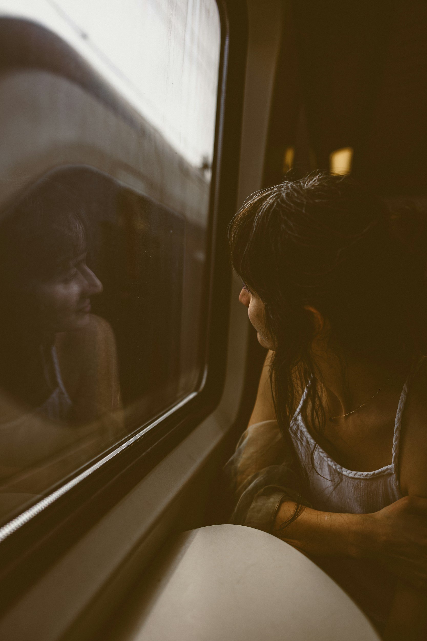A woman with dark hair looking out a window, her reflection visible on the glass.