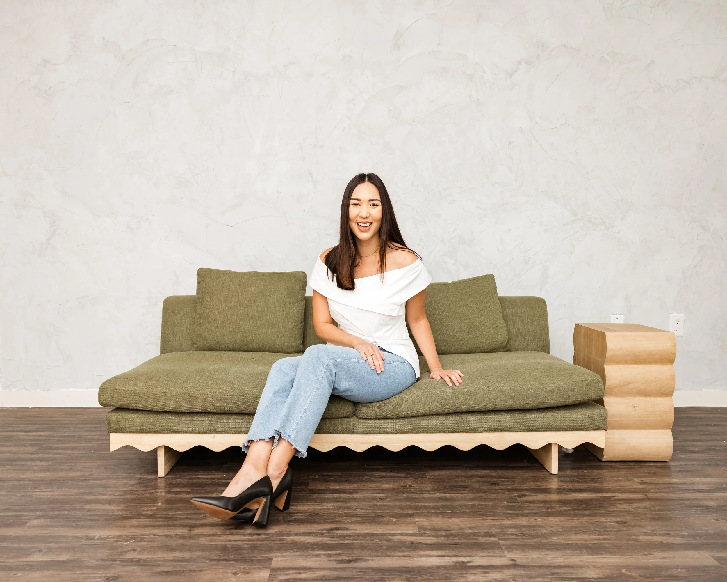 A smiling woman sitting on a green sofa with a white wall in the background.