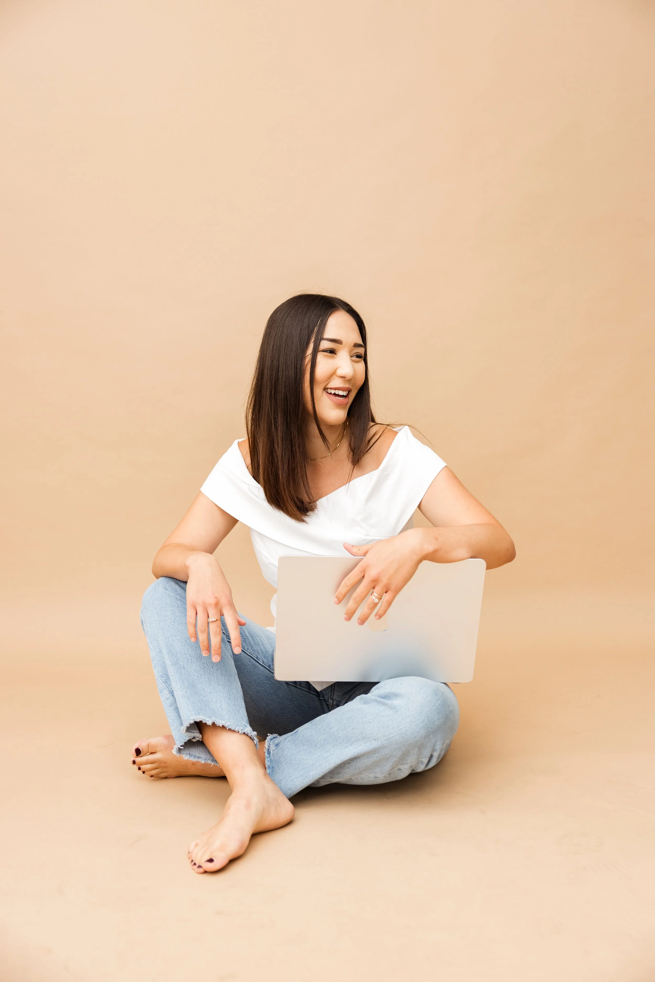 A woman sitting cross-legged on the floor, holding a laptop, smiling and looking to her right, wearing a white top and ripped jeans.